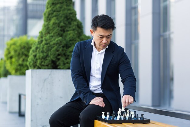 Uomo asiatico serio che gioca a scacchi fuori, uomo d'affari che pensa di giocare a scacchi seduto su una panchina vicino al centro dell'ufficio
