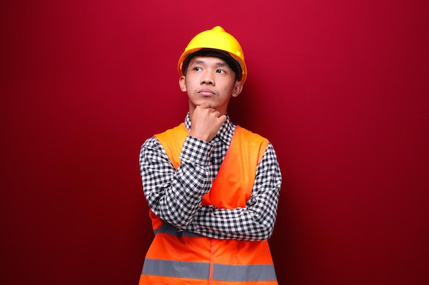 Uomo asiatico su sfondo rosso che indossa l'uniforme dell'appaltatore e il casco di sicurezza con l'espressione pensante