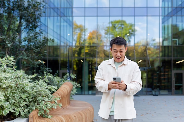 Uomo asiatico utilizzando il telefono al di fuori dell'edificio per uffici imprenditore libero professionista digitando il messaggio e sorridente