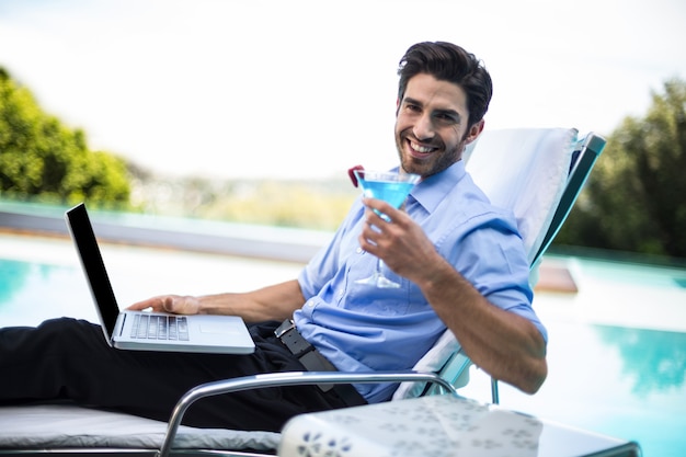 Uomo astuto che mangia martini mentre per mezzo del computer portatile vicino allo stagno