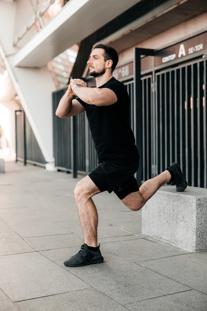 Uomo atletico che allunga le gambe prima dell'allenamento all'aperto. Vita attiva. concetto di sport urbano. Corridore in abbigliamento sportivo nero che si esercita al mattino tenendosi per mano nella serratura. Mattina riscaldamento.