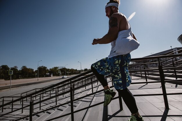 Uomo atletico con fascia in testa vestito con maglietta bianca, leggings neri e pantaloncini blu sta correndo giù per le scale fuori in una giornata di sole.