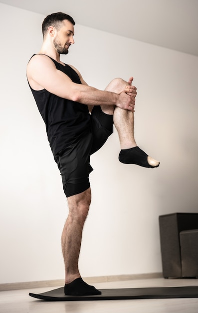 Uomo atletico in piedi su una gamba e allungandone un'altra. Allenamento sul tappetino yoga nero. Giovane muscolare che pratica posizione yoga a casa. Mattina riscaldamento. Abbigliamento sportivo nero. Stanza luminosa sullo sfondo.