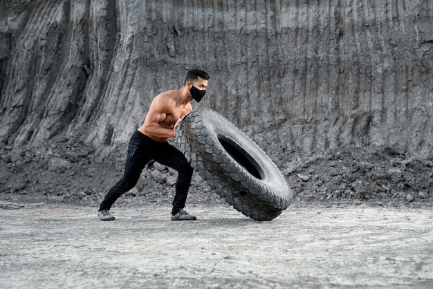 Uomo atletico senza camicia in maschera nera medica che spinge grande pneumatico pesante alla sabbiera. Concetto di bodybuilding e allenamento all'aperto.