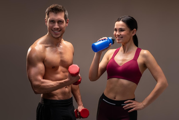 Uomo atletico sorridente che tiene in mano un manubrio rosso e guarda la telecamera mentre la donna beve acqua nelle vicinanze Studio indoor girato isolato su sfondo marrone