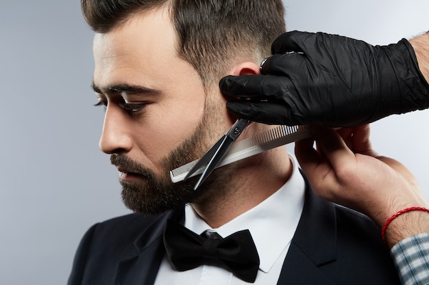 Uomo attraente al negozio di barbiere, guardando verso il basso, le mani dell'uomo che indossa la camicia che fa una forma di barba per l'uomo con i capelli neri al fondo dello studio, ritratto.