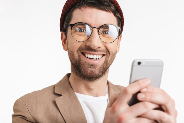 uomo attraente che indossa cappello e occhiali sorride mentre utilizza lo smartphone isolato su muro bianco