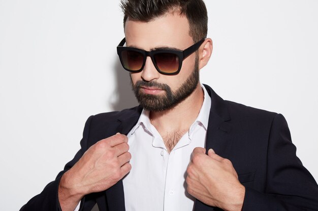 Uomo attraente con barba e capelli neri che indossa camicia bianca con cravatta, abito nero e occhiali da sole a sfondo bianco studio, ritratto, in posa, brutale.