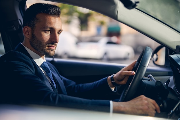 Uomo attraente in abiti formali con l'orologio a portata di mano che rimane concentrato mentre guida l'auto