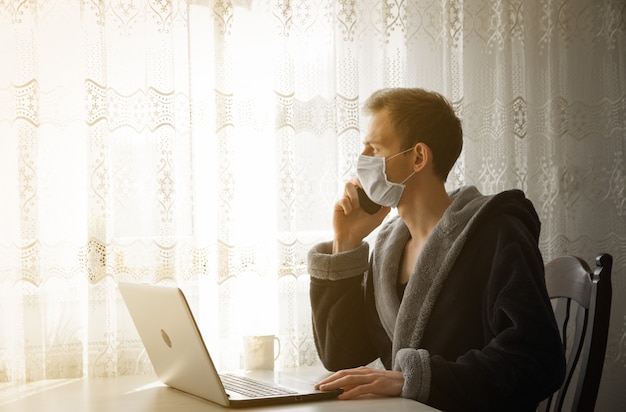 Uomo attraente in maschera medica durante la quarantena. Un giovane in un caffè lavora a un computer portatile vicino alla finestra. Isolamento, coronavirus, lavoro a casa.
