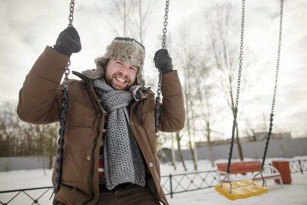 Uomo barbuto adulto in un cappello di pelliccia che oscilla su un'altalena per bambini nel parco in inverno