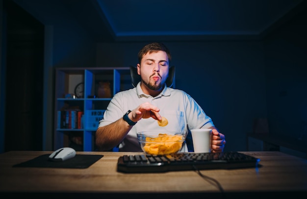 Uomo barbuto affamato che mangia patatine da un piatto di notte al computer a casa in una stanza
