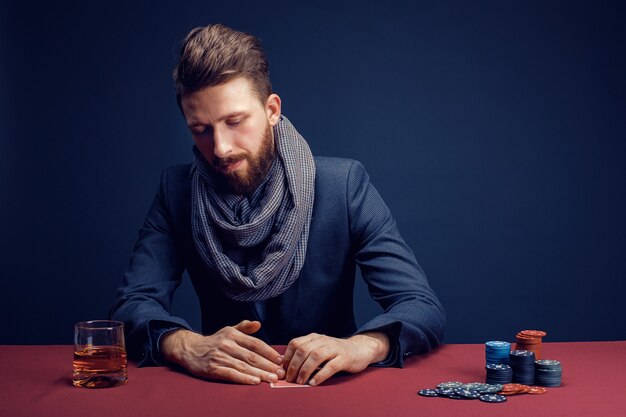 Uomo barbuto alla moda che gioca nel casinò scuro e beve whisky