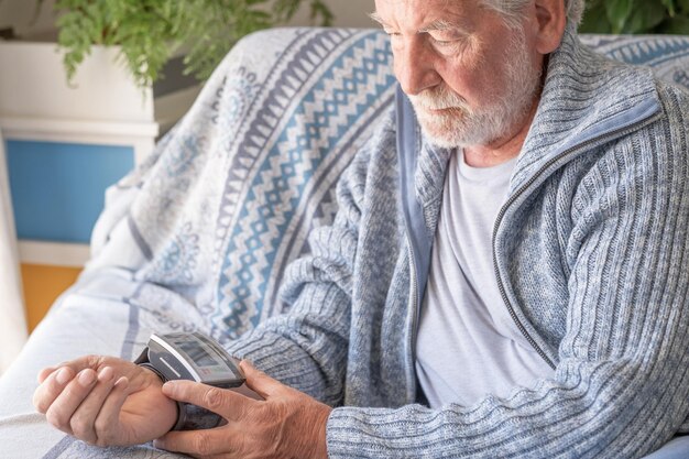 Uomo barbuto anziano che utilizza un dispositivo medico per misurare la pressione sanguigna nonno anziano seduto a casa sul divano