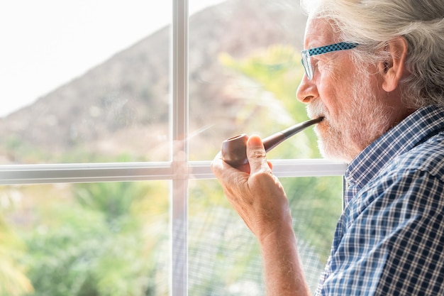 Uomo barbuto anziano pensieroso da solo all'interno che guarda fuori dalla finestra mentre fuma una pipa Concetto di anziani e di solitudine