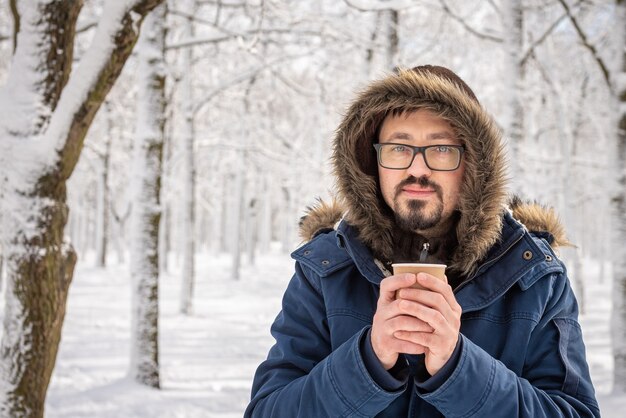 Uomo barbuto attraente in occhiali in posa all'aperto nel freddo dell'inverno. Ritratto di strada invernale. Caffè o tè in un bicchiere di carta nelle sue mani. Un uomo che si rilassa sulla passeggiata invernale nella foresta innevata.