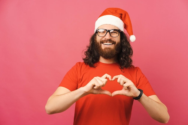 Uomo barbuto bello che indossa occhiali e cappello di Natale mostra il gesto del cuore