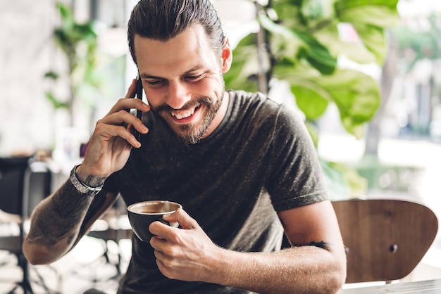 Uomo barbuto bello hipster utilizza smartphone con caffè al tavolo nella caffetteria