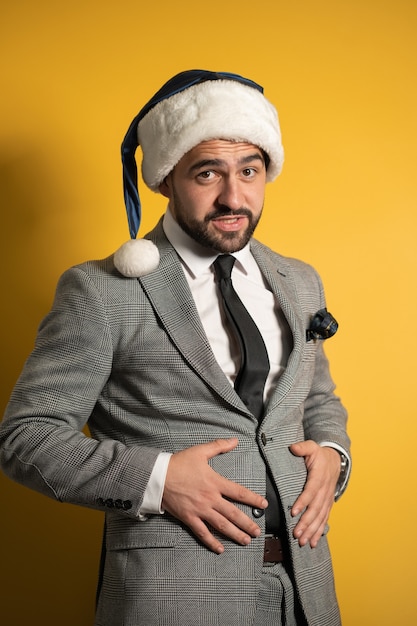 Uomo barbuto bello in cappello blu della Santa e vestito grigio, guardando la parte anteriore isolata sul muro giallo