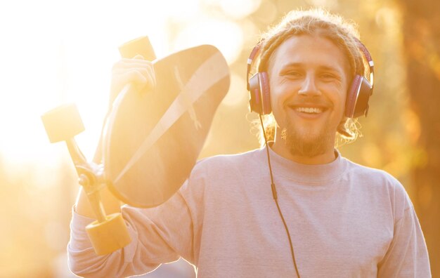 Uomo barbuto biondo skater hipster con dreadlocks che indossa le cuffie ascoltando la sua musica preferita e ridendo al tramonto nella vista all'aperto della foresta