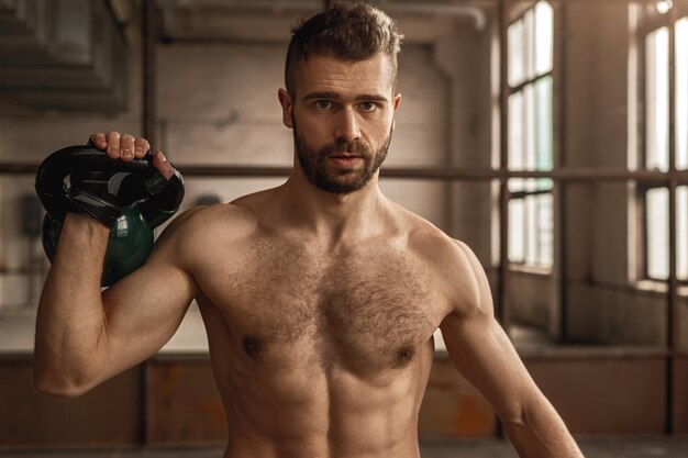 Uomo barbuto brutale fiducioso con torso muscoloso nudo che fa esercizio con kettlebell pesante durante un intenso allenamento in palestra