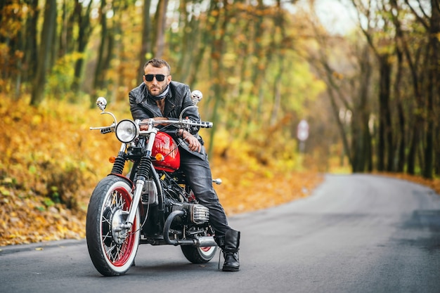 Uomo barbuto brutale in occhiali da sole e giacca di pelle seduto su una moto sulla strada nella foresta