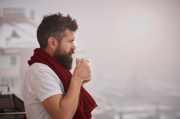 Uomo barbuto brutale in piedi fuori con un asciugamano rosso sulle spalle, tenendo la tazza con una bevanda calda fumante. Giovane uomo spensierato bello che gode di una tazza di caffè mentre guarda fuori, buongiorno.