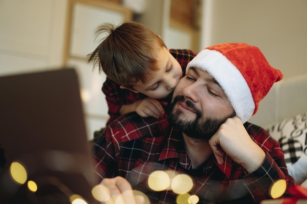 Uomo barbuto caucasico in camicia di flanella scozzese rossa con cappello da Babbo Natale con il suo piccolo figlio carino che lo abbraccia