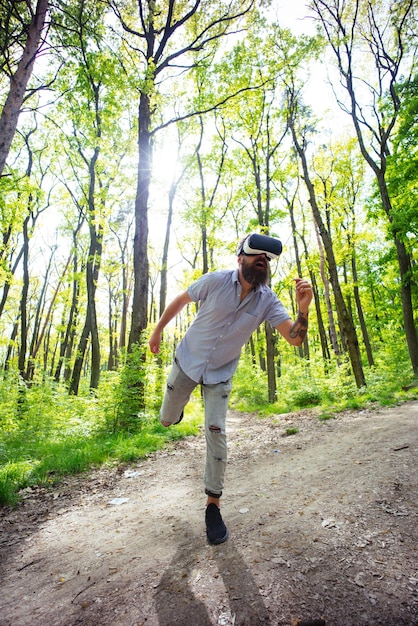 Uomo barbuto che ha esperienza 3D con gli occhiali VR. Hipster emozionante che prova il nuovo gadget nella foresta. Uomo che corre nella realtà virtuale, concetto di era digitale