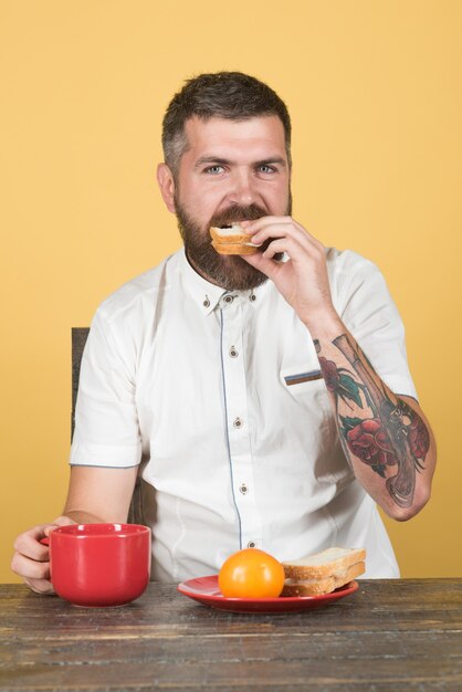 Uomo barbuto che mangia colazione con una tazza di caffè mattutino e toast al pomodoro in mano sul piatto tazza