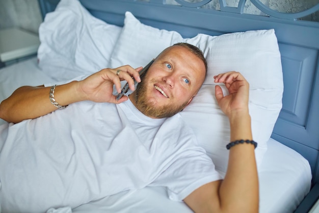 Uomo barbuto che parla smartphone mentre è sdraiato su un letto accogliente