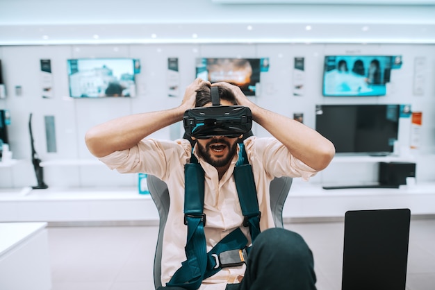 uomo barbuto che prova la tecnologia della realtà virtuale mentre è seduto sulla sedia nel negozio di tecnologia.