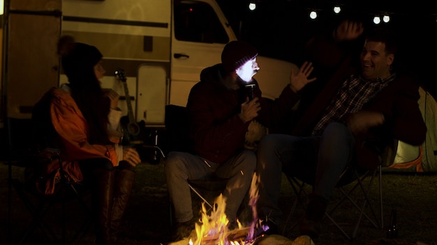 Uomo barbuto che racconta una storia spaventosa ai suoi amici intorno al fuoco da campo in una fredda notte d'autunno. Camper retrò. Lampadine