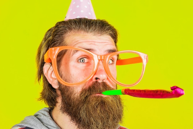 Uomo barbuto che soffia il ragazzo del tempo della festa di compleanno del noisemaker con cappello a cono e occhiali grandi