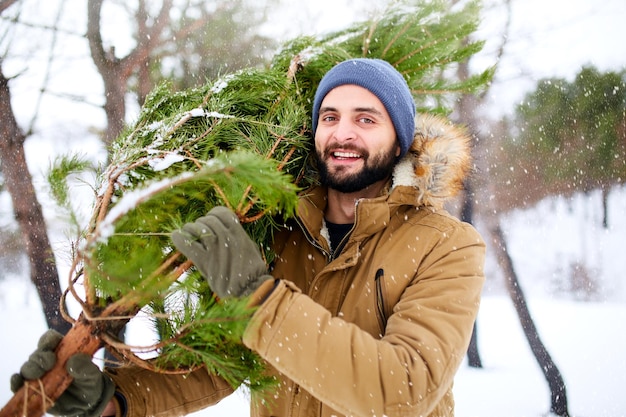 Uomo barbuto che trasporta albero di natale appena tagliato nella foresta il giovane boscaiolo porta l'albero di abete