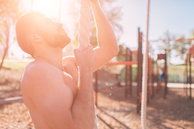 Uomo barbuto con capelli neri arrampicata su corda spessa durante esercizi di fitness in allenamento cross-fit con