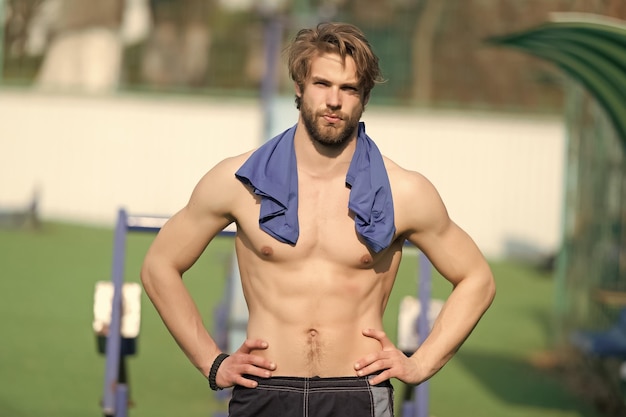 Uomo barbuto con muscoli del busto in forma allo stadio Sportivo con maglietta blu sul collo Atleta con pancia e petto sexy all'aperto soleggiato Allenamento e allenamento Sport o fitness e cura del corpo