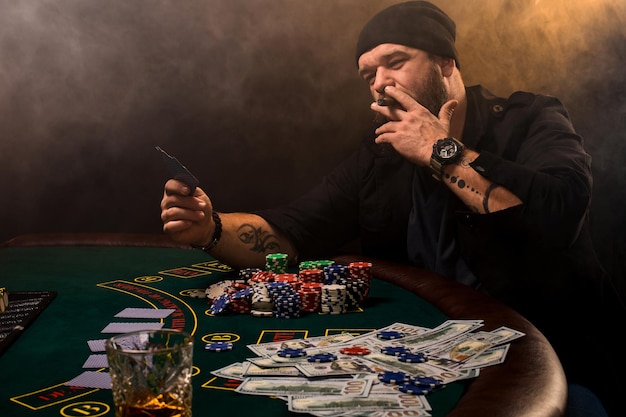 Uomo barbuto con sigaro e bicchiere seduto al tavolo da poker in un casinò. Gioco d'azzardo, carte da gioco e roulette.