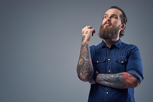 Uomo barbuto con tatuaggi sulle braccia, vestito con una camicia di jeans su sfondo grigio.