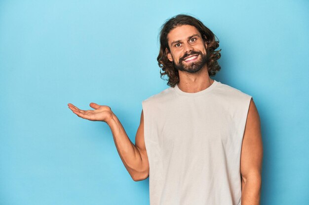 Uomo barbuto con un tank top sullo sfondo blu che mostra uno spazio di copia su un palmo e che tiene un'altra mano