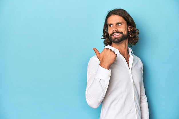 Uomo barbuto con una camicia bianca sullo sfondo blu punta con il pollice lontano ridendo e spensierato