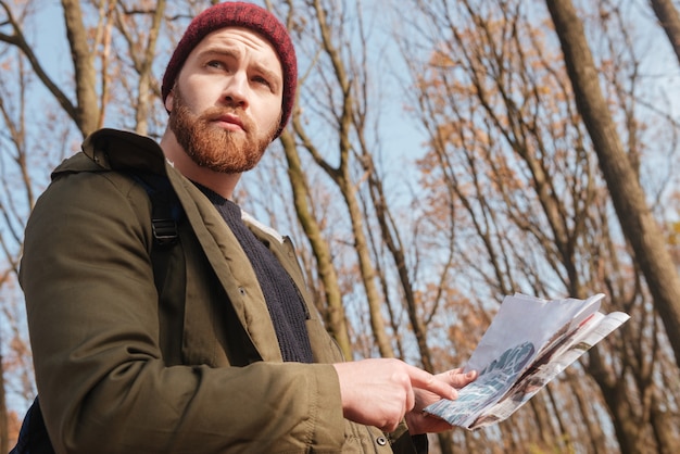 Uomo barbuto concentrato che indossa un cappello che tiene la mappa nelle mani in piedi nella foresta