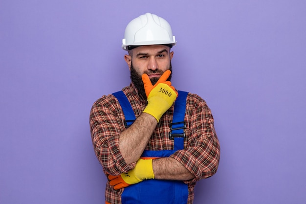 Uomo barbuto costruttore in uniforme da costruzione e casco di sicurezza che indossa guanti di gomma guardando con la mano sul mento pensando