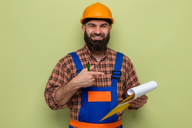 Uomo barbuto costruttore in uniforme da costruzione e casco di sicurezza che tiene appunti che puntano con il dito indice sorridendo allegramente in piedi su sfondo verde