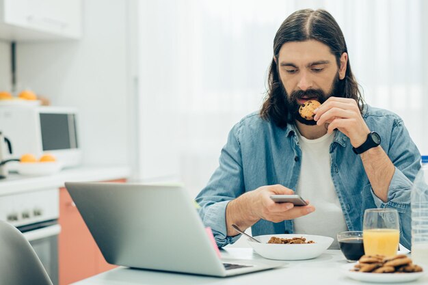 Uomo barbuto dai capelli lunghi che morde un biscotto al cioccolato e guarda attentamente lo schermo del suo smartphone