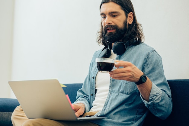 Uomo barbuto dai capelli lunghi concentrato seduto davanti allo schermo di un laptop e con in mano una tazza di caffè