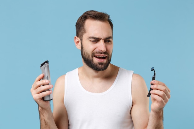 Uomo barbuto di 20 anni in camicia bianca tenere scelta rasoio di plastica elettrico isolato su sfondo blu pastello ritratto in studio. Concetto di procedure cosmetiche sanitarie per la cura della pelle. Simula lo spazio della copia.