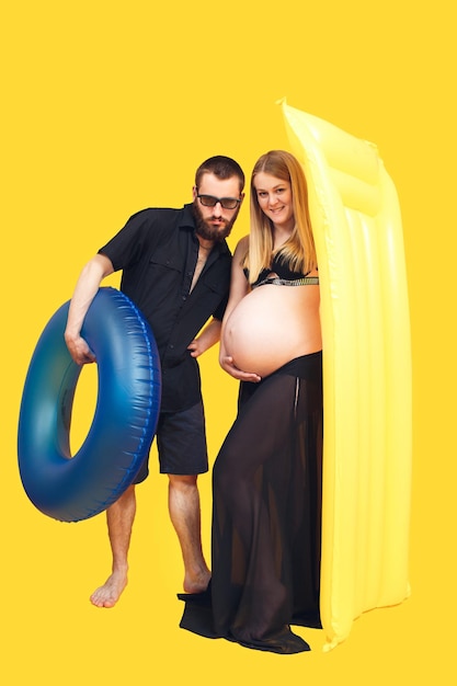 Uomo barbuto e donna incinta che tengono anello gonfiabile e materasso su sfondo giallo