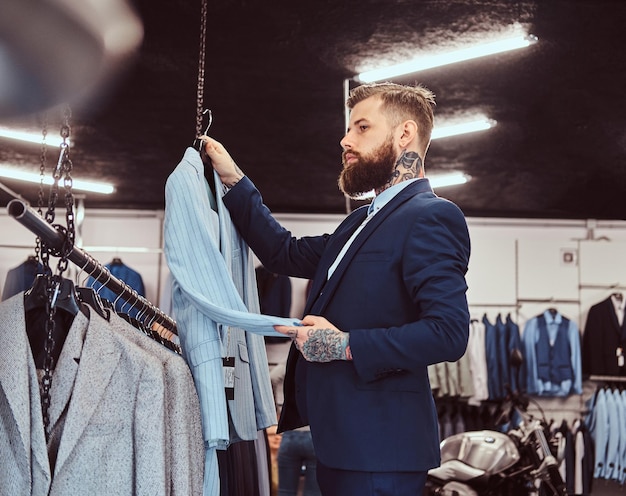 Uomo barbuto elegantemente vestito con tatuaggi su mani e collo sceglie un nuovo abito nel negozio di abbigliamento maschile.