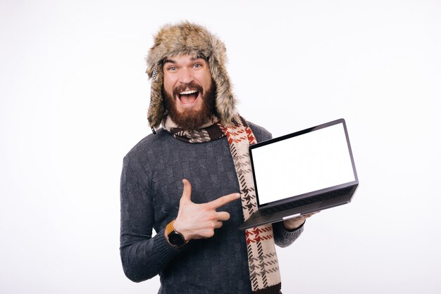 Uomo barbuto emozionante che indossa cappello o berretto caldo invernale, una sciarpa è imbronciata al suo portatile schermo bianco vuoto.